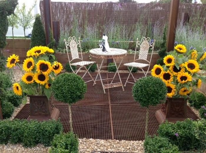 A small bistro table set among standard topiary and sunflowers on a brick path