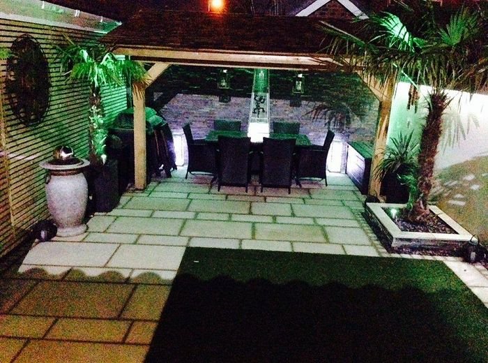A lit pergola over a paved seating area with planting either side