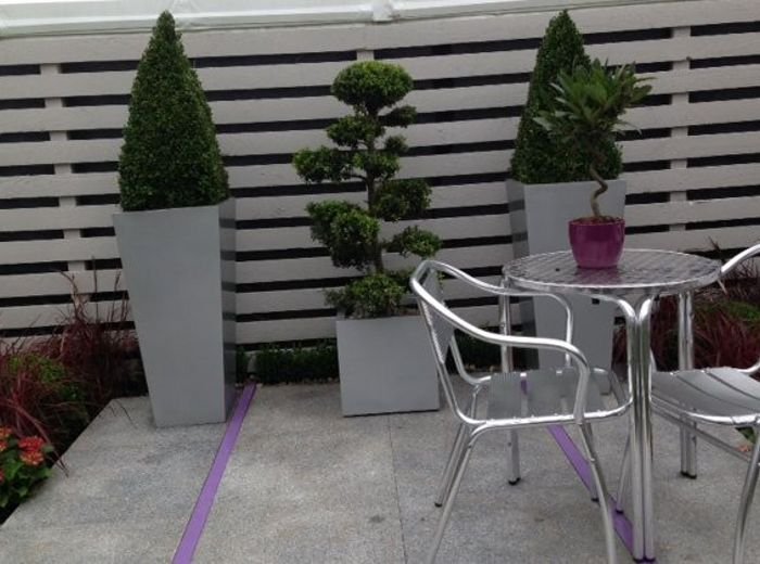 Tall grey planters holding clipped topiary beside a paved terrace and café table