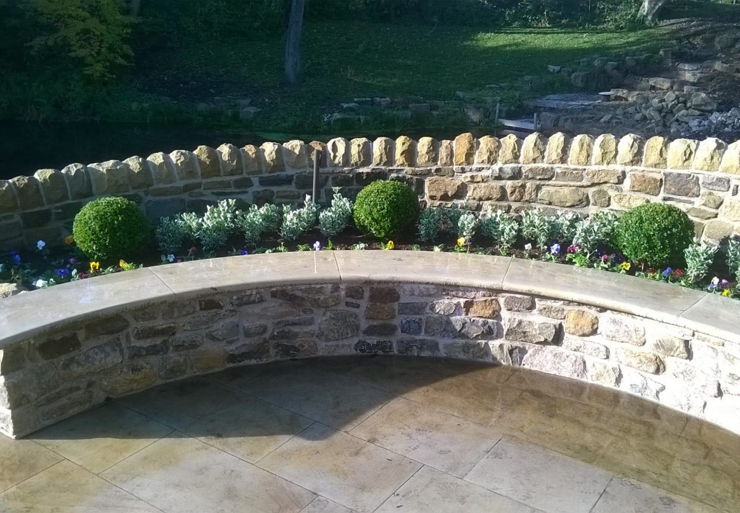 A curved stone seating wall with clipped box balls and spring bedding above a flagged terrace, built by New Leaf